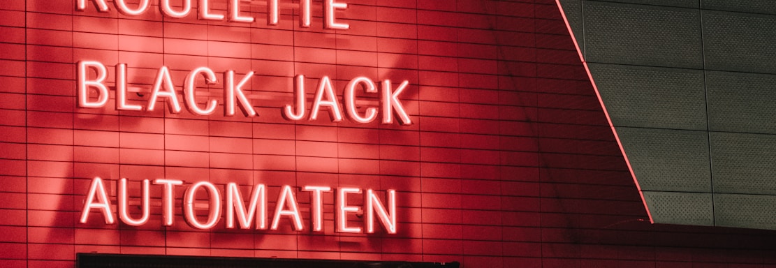 Red neon letters are mounted on a dark tiled wall, displaying the words Poker, Sic Bo, Roulette, Black Jack, and Automaten.