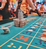 A casino scene with a roulette table features prominently, covered in green felt with colorful numbers and betting squares. Several stacks of poker chips are placed on different numbers, and the hands of several people are visible as they interact with the table. In the background, there are slot machines and more people, creating a vibrant and busy atmosphere.