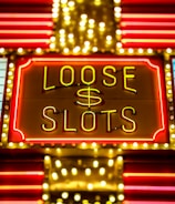 Bright neon lights display the words 'Loose Slots' with a dollar sign in the center. The sign is outlined with bulbs, creating a vibrant and flashy effect typical of a casino or entertainment venue.