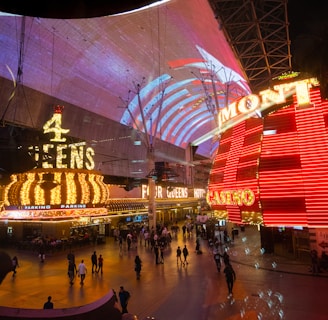 A bustling nighttime scene with illuminated casino signage towering over a busy pedestrian street. Neon lights in red and yellow highlight the names of various establishments. People are walking in different directions, enjoying the vibrant atmosphere.