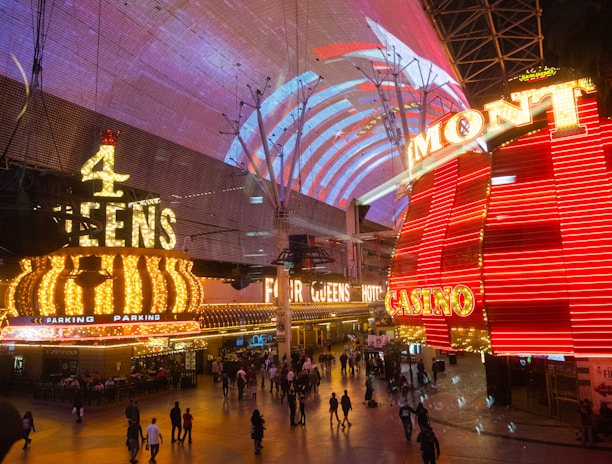 A bustling nighttime scene with illuminated casino signage towering over a busy pedestrian street. Neon lights in red and yellow highlight the names of various establishments. People are walking in different directions, enjoying the vibrant atmosphere.