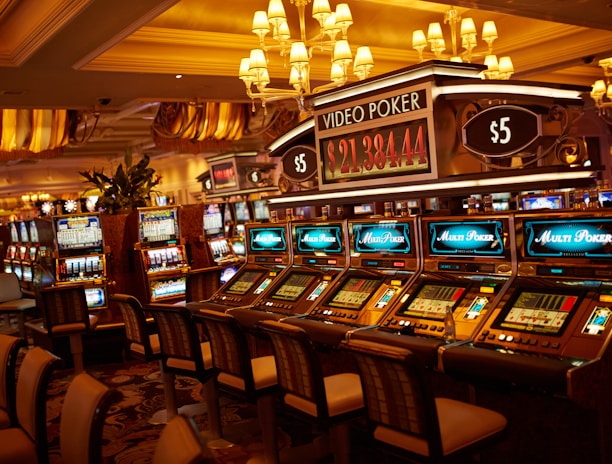 A casino floor featuring rows of brightly lit video poker machines. The area is illuminated by elegant chandeliers, with a display board showing a large sum of money, suggesting a possible jackpot. The rich decor includes luxurious carpets and comfortable seating, adding to the opulent atmosphere.
