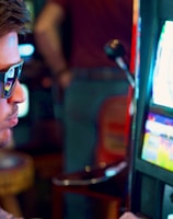 A man wearing sunglasses is intently focused on a slot machine, with colorful lights and graphics illuminating the screen. The scene is set indoors, likely in a casino or gaming area. His attire includes a patterned shirt and a blazer.