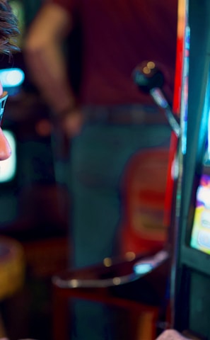 A man wearing sunglasses is intently focused on a slot machine, with colorful lights and graphics illuminating the screen. The scene is set indoors, likely in a casino or gaming area. His attire includes a patterned shirt and a blazer.