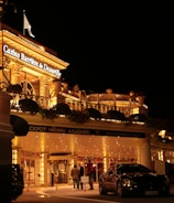 A luxurious casino at night, illuminated with warm, golden lights. The building features elegant architecture with columns and decorative elements. A few people are walking towards the entrance, and there are a couple of parked cars nearby.