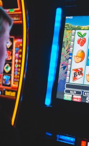 A person is closely observing a digital slot machine with colorful fruit symbols, bars, and a bell. The slot machine screen displays symbols like oranges, plums, cherries, and clouds. The background is dimly lit, creating a focused and somewhat intense atmosphere.