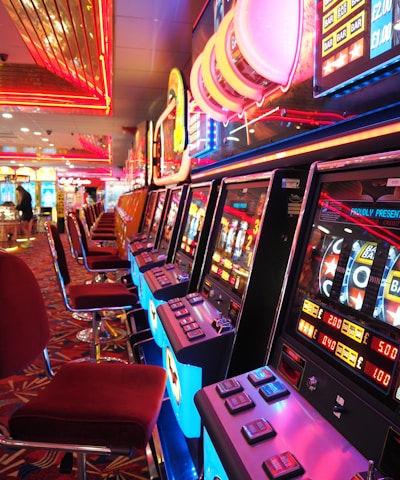 A brightly lit casino interior featuring a row of colorful slot machines. The neon lights create a vibrant atmosphere, with patterned carpeting adding to the decor. Stools are aligned neatly in front of each machine.