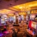 A vibrant casino interior filled with rows of colorful slot machines, each illuminated with bright lights and displays. The ceiling is adorned with elegant chandeliers and golden drapes, creating a luxurious atmosphere. The patterned carpet adds to the elaborate design of the space.