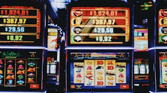 A row of colorful slot machines lit with bright neon lights, showcasing various graphical elements on the screens, indicating different games and jackpots. The atmosphere suggests a lively casino setting, bustling with excitement.
