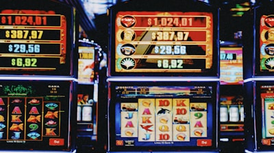 A row of colorful slot machines lit with bright neon lights, showcasing various graphical elements on the screens, indicating different games and jackpots. The atmosphere suggests a lively casino setting, bustling with excitement.