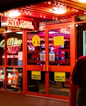 A brightly lit entrance to a casino with neon signs, including one advertising a £500 jackpot. The doors have clear signage indicating entry is restricted to those over 18. A person in a red jacket stands to the right, possibly interacting with a machine or display.