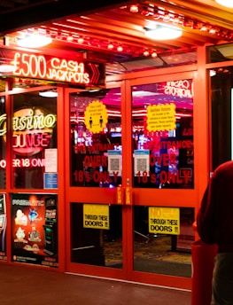 A brightly lit entrance to a casino with neon signs, including one advertising a £500 jackpot. The doors have clear signage indicating entry is restricted to those over 18. A person in a red jacket stands to the right, possibly interacting with a machine or display.
