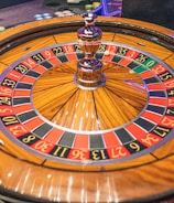 A roulette wheel with alternating red and black numbered pockets on a wooden platform. The wheel contains a small white ball resting in one of the pockets, indicating gameplay. Surrounding the wheel are stacks of colorful poker chips, suggesting a casino environment.