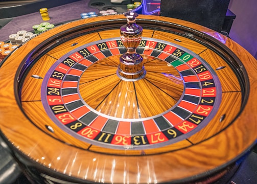 A roulette wheel with alternating red and black numbered pockets on a wooden platform. The wheel contains a small white ball resting in one of the pockets, indicating gameplay. Surrounding the wheel are stacks of colorful poker chips, suggesting a casino environment.