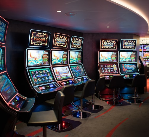 A row of brightly lit slot machines in a casino setting, each with colorful displays and various game themes. The machines are lined up against a dark wall and are unoccupied, with cushioned chairs placed in front of them. The area is dimly lit except for the glow from the screens, creating a vibrant and engaging atmosphere.