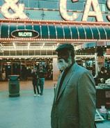 An older man with a gray beard and wearing a suit and hat stands in front of a brightly lit casino. The background features a large illuminated sign reading 'CASINO' and 'SLOTS' among other words, with numerous lights outlining the entrance. Several other people are visible in the background, engaged in various activities, some walking, some standing, and one looking at something on display.