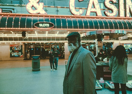 An older man with a gray beard and wearing a suit and hat stands in front of a brightly lit casino. The background features a large illuminated sign reading 'CASINO' and 'SLOTS' among other words, with numerous lights outlining the entrance. Several other people are visible in the background, engaged in various activities, some walking, some standing, and one looking at something on display.