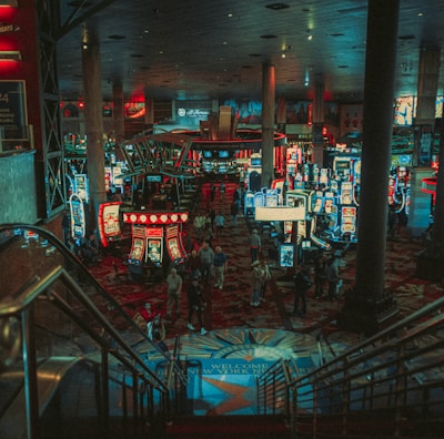 A bustling casino floor filled with groups of slot machines and people walking around. The atmosphere is vibrant with neon lights and illuminated signs. The interior has a ceiling supported by large pillars and there are stairs leading down to the gaming area.